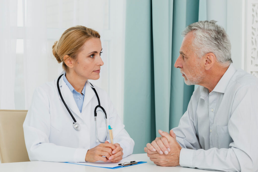 A medical professional, identifiable by a white coat and stethoscope, sits at a desk and converses with an older individual. The medical professional is holding a pen and taking notes on a clipboard. The older individual, with hands clasped together, suggests a consultation or discussion in a medical office or clinic setting.