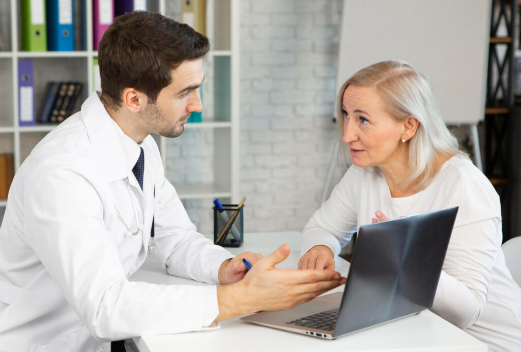 A doctor in a white coat is holding a pen and speaking with a patient, who has a laptop open in front of them. The setting is an office environment with shelves containing colorful binders and books in the background, suggesting a detailed discussion about the patient's health or medical records.