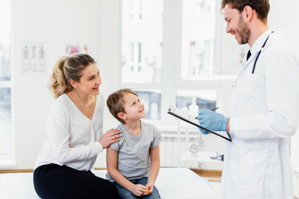 A healthcare professional, dressed in a white coat and wearing gloves, is holding a clipboard and speaking with a young child who is sitting on an examination table. An adult is seated next to the child, with a hand placed on the child's shoulder. The room is well-lit with large windows, and medical posters are visible on the wall.