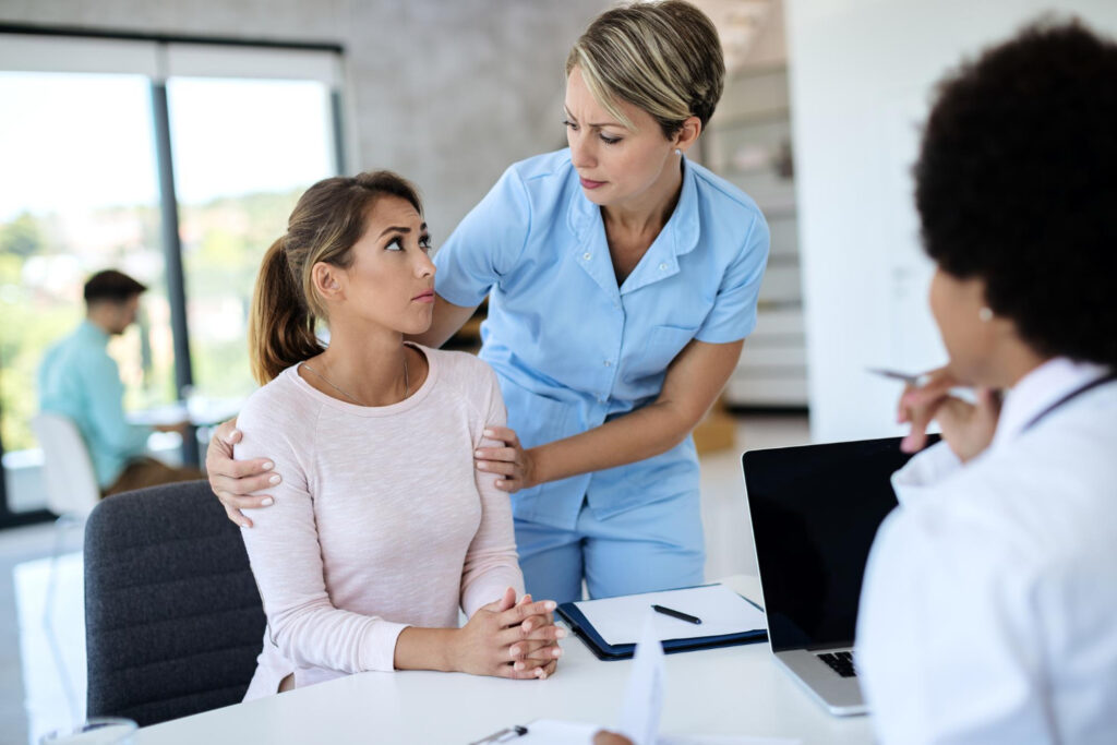 A healthcare professional in a blue uniform is comforting a patient at Calgary Medical Center & Walk in Clinic. Another healthcare professional is seated at a desk with a laptop and documents, engaging with the patient.